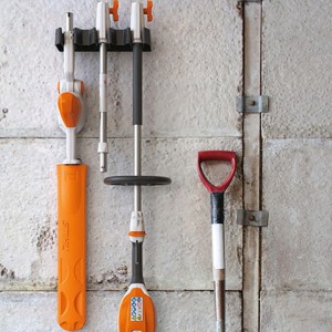 Stihl HLA 56 Nożyce akumulatorowe, na wysięgniku bez akumulatora i bez ładowarki, zawieszone na ścianie obok łopaty.